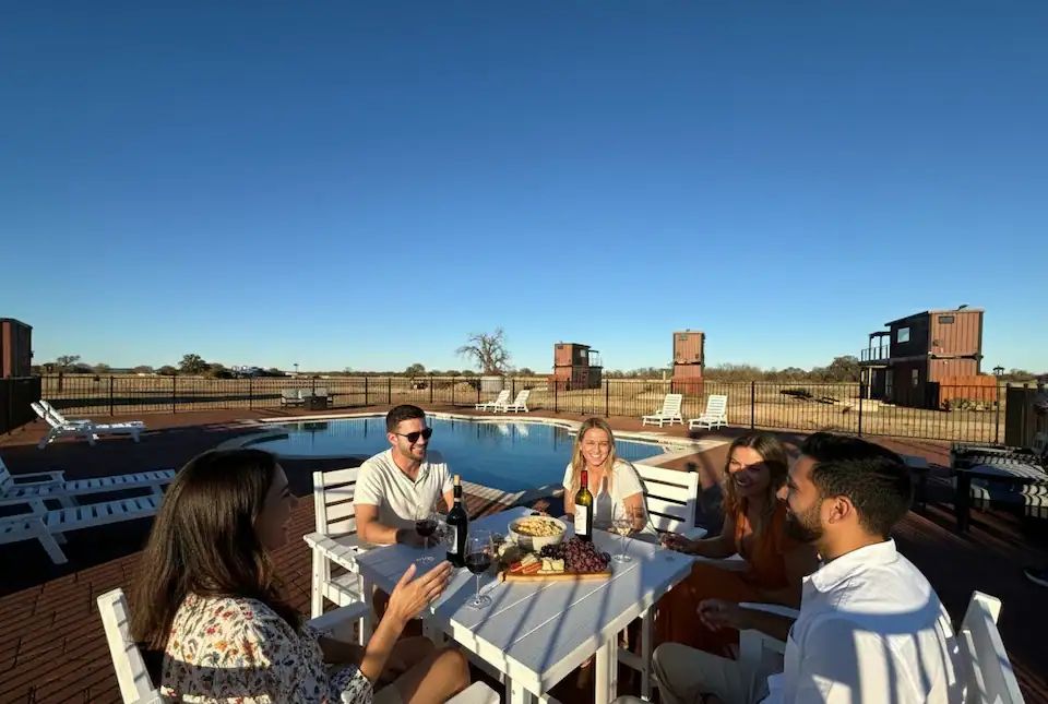 A man and woman sitting across from each other at a modern fire pit table on a wooden rooftop deck during sunset.